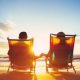 A man and woman facing the sunset while relaxing in beach chairs and holding hands