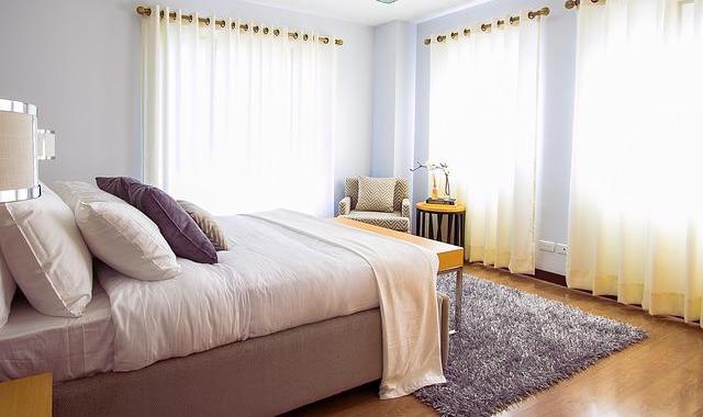 A tastefully decorated bedroom with a queen-sized bed. Three large windows have sheer curtains that let in the light. A shag area rug is under the bed.